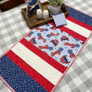 Könnte beinhalten: Ein roter, weißer und blauer gesteppter Tischläufer mit patriotischem Thema. Der Läufer zeigt ein blau-weißes Gingham-Muster, rote Lastwagen und Sterne. Der Läufer ist mit einem weißen und roten Rand eingefasst.