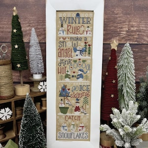 Puede incluir: Un muestrario de punto de cruz enmarcado verticalmente sobre tela color canela con el texto WINTER RULES, make a snow angel, drink hot cocoa, go ice skating y CATCH SNOWFLAKES, acompañado de coloridas ilustraciones bordadas de muñecos de nieve.