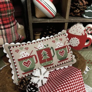 Könnte beinhalten: Ein besticktes Kissen mit Weihnachtstassen, Tannenbäumen und Herzen. Es ist umgeben von rot-weißem Vichystoff, Tannenzapfen und einer Holzschneeflocke. Der Text "Cup of Cheer" und "Primrose Cottage" sind sichtbar.
