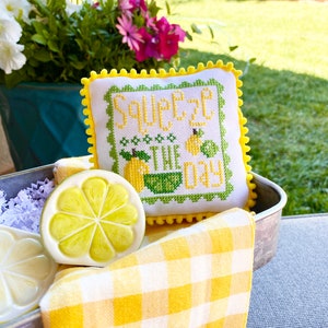 May include: A yellow and white gingham fabric with a yellow lemon slice on top. A small white pillow with yellow trim and a cross-stitch design that reads "Squeeze the Day" is in the background.