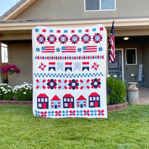 May include: A red, white, and blue quilt with a patriotic theme. The quilt features stars, stripes, and houses. The quilt is hanging on a wall outside a house.