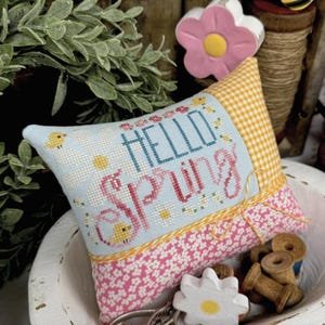 Puede incluir: Un cojín decorativo con la frase "Hello Spring" bordada en azul y rosa, con motivos florales y de pájaros. El cojín tiene un borde con estampado floral rosa y una parte superior a cuadros amarillos. La imagen también incluye un cuenco de madera y elementos decorativos.