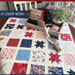 Może przedstawiać: Kołdra w kolorze czerwonym, białym i niebieskim z wzorem gwiazd. Kołdra ma wymiary 86 cm x 86 cm i nosi nazwę "Falling Stars" autorstwa Lindsey Weight. Kołdra jest wykonana z różnych tkanin, w tym wzorów kwiatowych, kropek i jednokolorowych.