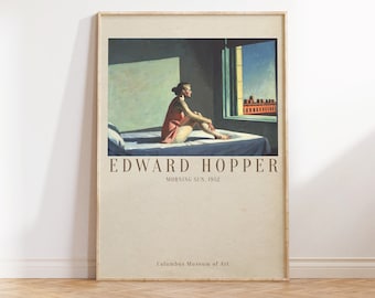 May include: A painting by Edward Hopper titled "Morning Sun" from 1952. The painting depicts a woman sitting on a bed in a room with a window overlooking a city street. The woman is wearing a pink dress and is looking out the window. The painting is in a light brown frame.