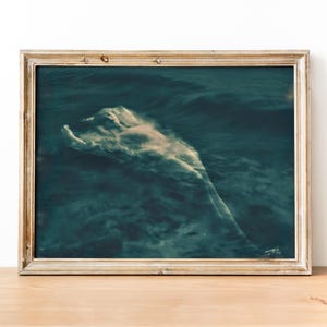 May include: A framed art print featuring a serene, ethereal image of a figure submerged in dark blue water. The figure appears as a soft, light-colored form amidst the waves, creating a dreamlike atmosphere. The frame is a light, neutral color.