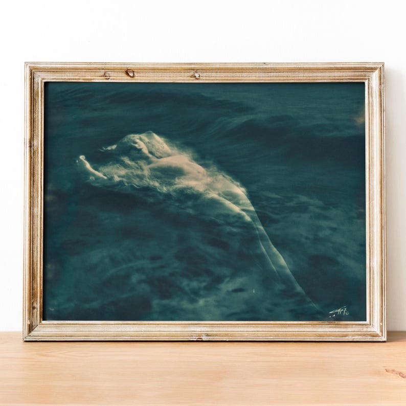 May include: A framed art print featuring a serene, ethereal image of a figure submerged in dark blue water. The figure appears as a soft, light-colored form amidst the waves, creating a dreamlike atmosphere. The frame is a light, neutral color.