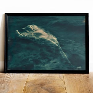 May include: A framed art print featuring a figure submerged in dark teal water. The figure is partially visible, with a cloud-like effect around the body. The artwork is framed in a black border and rests on a wooden surface.