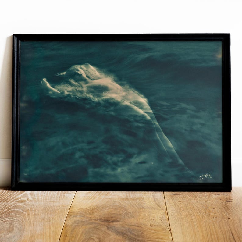 May include: A framed art print featuring a figure submerged in dark teal water. The figure is partially visible, with a cloud-like effect around the body. The artwork is framed in a black border and rests on a wooden surface.