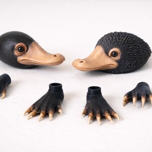 May include: Two black and tan Niffler heads with detailed eyes and beaks, along with four black and tan feet with claws. The items are arranged on a white surface, suggesting a craft or collectible theme.