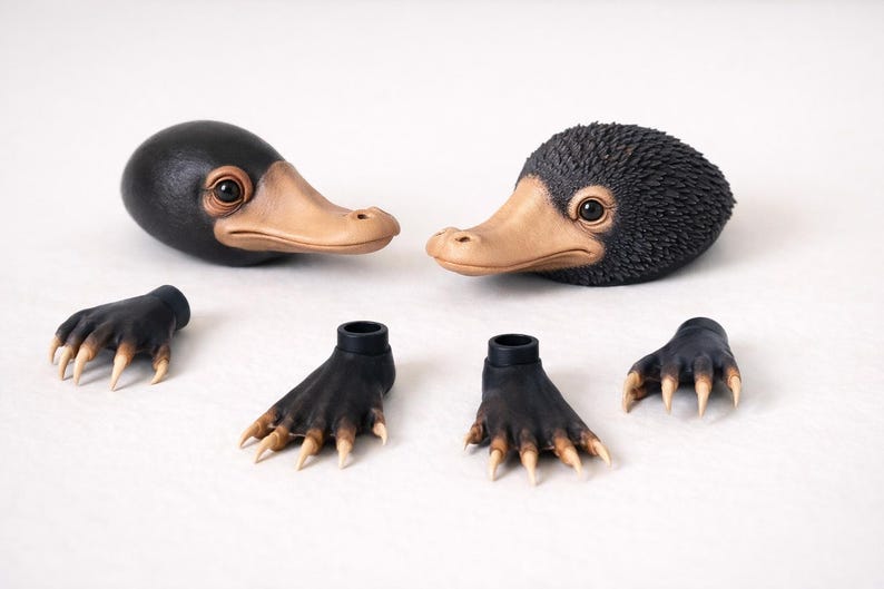 May include: Two black and tan Niffler heads with detailed eyes and beaks, along with four black and tan feet with claws. The items are arranged on a white surface, suggesting a craft or collectible theme.