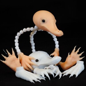 May include: A disassembled, whimsical creature sculpture with a light peach-colored head and beak, white body, and articulated limbs. The sculpture is set against a black background, highlighting its unique design.