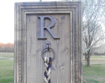 May include: A wooden plaque with a brown frame and a metal ring. The plaque has a large metal letter "R" and a braided rope hanging from the ring. The plaque has the text "A cord of three strands is not quickly broken Ecclesiastes 4:12" written on it.