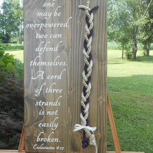 Pode incluir: Uma placa de madeira com uma inscrição branca que diz "Though one may be overpowered, two can defend themselves. A cord of three strands is not easily broken. Ecclesiastes 4:12". Uma corda trançada com três fios em roxo, dourado e branco está presa à placa.