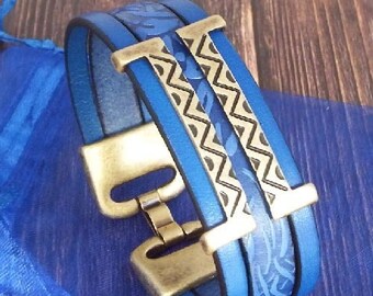 May include: A blue leather bracelet with a gold metal clasp and a geometric pattern. The bracelet has a woven design and a decorative metal accent.