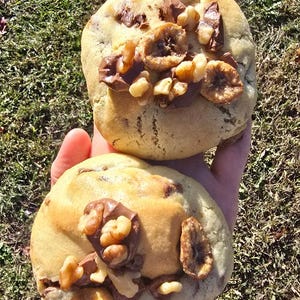 May include: Two large chocolate chip cookies with walnuts and chocolate chunks on top. The cookies are held in a hand.