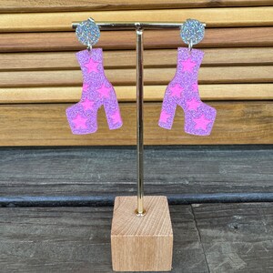May include: A pair of pink platform shoe earrings with silver glitter and star accents. The earrings are displayed on a wooden stand.