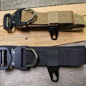 Puede incluir: Se muestran dos cinturones tácticos, uno en color arena y otro en negro. Ambos tienen una hebilla metálica con anilla en D, un gancho y una correa de velcro. Los cinturones están hechos de cincha de nailon duradera, aptos para actividades al aire libre.