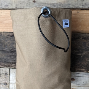 Könnte beinhalten: Beigefarbener Canvas-Beutel mit schwarzem Kordelzug und Metallöse. Der Beutel hat ein kleines weißes Etikett mit dem Text "ON POINT GEAR" und einer Bär-Grafik. Der Hintergrund ist aus Holz.