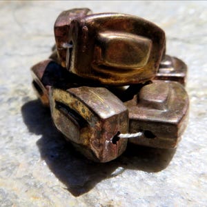 Puede incluir: Un montón de cuentas de color marrón metálico con forma rectangular y una parte superior ligeramente curvada. Cada cuenta tiene un agujero para ensartar. Las cuentas tienen un aspecto vintage y desgastado, con toques de oro y bronce.