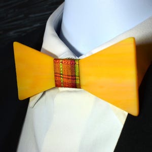 May include: A bow tie featuring two solid, smooth, yellow, trapezoidal-shaped panels connected by a central fabric band. The central band displays a multi-colored plaid pattern in shades of orange, red, and green. The bow tie is worn on a white shirt.
