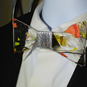 Könnte beinhalten: Eine einzigartige Fliege mit einem transparenten, geometrischen Design. Die Fliege hat eine silberne Mitte und ist mit farbenfrohen abstrakten Formen in Orange, Gelb und Schwarz verziert. Sie wird mit einem weißen Hemd mit Kragen und einem schwarzen Anzug getragen.