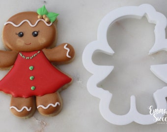 May include: Gingerbread girl cookie cutter with a gingerbread girl cookie decorated with red icing, green icing, and white icing. The cookie cutter is white plastic.