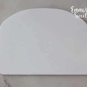 May include: White plastic scraper tool for smoothing out fondant or icing on cakes. It has a rounded top and a flat bottom.