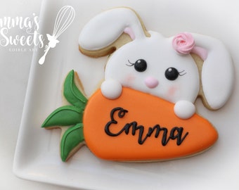 Puede incluir: Una galleta de conejito blanco sosteniendo una galleta de zanahoria con el nombre "Emma" escrito en glaseado negro. El conejito tiene mejillas rosadas, ojos negros y una flor rosa en la cabeza.