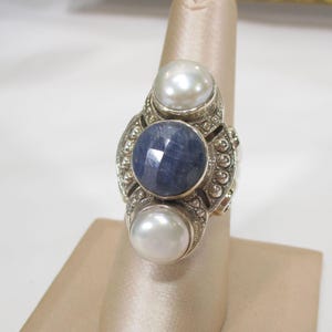 May include: A silver ring featuring a large, faceted, dark blue gemstone, flanked by two white pearls. The ring has an ornate, detailed setting with a vintage aesthetic. The ring is displayed on a neutral background.