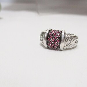 May include: A silver ring featuring a central band of small, round, deep pink stones set in a dark metal base. The sides of the ring are crafted from polished silver with a textured, diagonal, woven-like pattern that extends around the band.