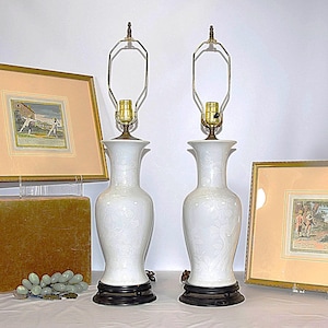 May include: Pair of white ceramic table lamps with floral patterns. Each lamp has a brass neck, a black base, and a metal lamp shade frame. Framed artwork and decorative objects are in the background.