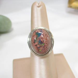 May include: A silver ring featuring an oval-shaped opal gemstone. The opal has a reddish-brown base color with iridescent flashes of green and blue. The gemstone is set within a double-wire silver band.