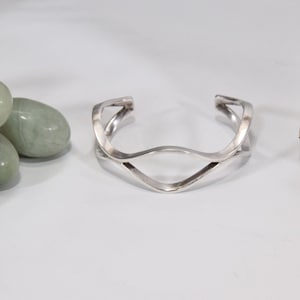 May include: A silver cuff bracelet with a unique, open design. The bracelet features a wavy, intertwined pattern, creating an eye-catching visual effect. The metal has a polished finish, reflecting light. The jewelry is displayed on a white surface.