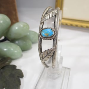 May include: Silver-toned cuff bracelet with a turquoise stone and leaf accents. The oval turquoise stone is set in a rope-like border. The bracelet features two leaf designs on the sides.