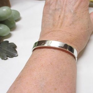 May include: A polished silver cuff bracelet. The bracelet is a simple, flat band with a reflective surface. The bracelet is worn on a wrist, showcasing its minimalist design and bright shine. Jewelry.
