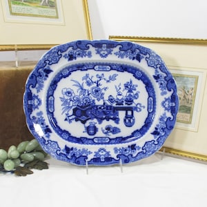 May include: Oval blue and white porcelain platter with a floral and landscape design. The platter has a scalloped edge and is supported by a clear stand. The design features a central scene with flowers, a table, and other decorative elements.