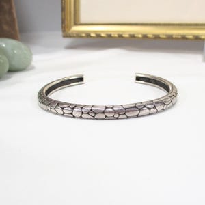 Peut inclure: Un bracelet manchette argenté avec un motif texturé en forme d'écailles. Le bracelet ouvert est présenté sur une surface blanche, avec un cadre doré en arrière-plan. Le design du bracelet lui donne un aspect unique et accrocheur.