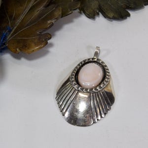 May include: A silver pendant with a light pink oval stone at the center. The pendant has a decorative border around the stone and a fan-like design on the bottom half. The pendant is on a white surface.
