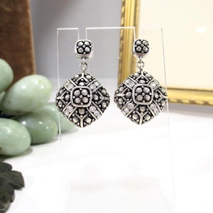 May include: A pair of silver-toned earrings with a square, ornate design. Each earring features a smaller square top and a larger, diamond-shaped pendant. The pendants are embellished with small, clear stones and a detailed pattern.
