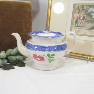 May include: A vintage white ceramic teapot with a blue speckled band and lid. The teapot features a red floral design with green leaves. It has a curved spout and a handle. The teapot is a classic piece of kitchenware.