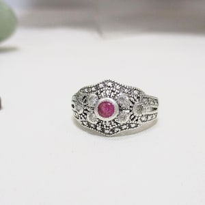 May include: A silver ring with a central round red gemstone, surrounded by smaller clear stones and intricate metalwork. The ring has a vintage or antique style, with a detailed, ornate design. The ring is displayed on a white surface.