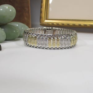 May include: A wide, flexible link bracelet featuring alternating vertical segments of polished silver-tone and textured gold-tone metal. The design consists of repeating rectangular panels with a woven, mesh-like appearance and a secure clasp.