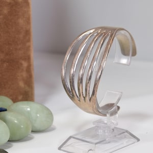 May include: Silver-toned cuff bracelet with a series of vertical bars. The open design allows light to pass through. The bracelet is displayed on a clear plastic stand, showcasing its elegant, minimalist style. The bracelet is a stylish accessory.