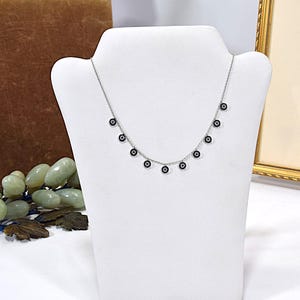 May include: A silver necklace with multiple small, circular charms. Each charm has a dark center surrounded by a lighter color, resembling an eye. The necklace is displayed on a white mannequin, showcasing its delicate design.