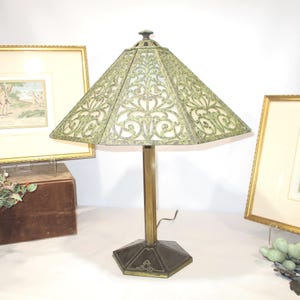 May include: An antique table lamp with a geometric shade featuring an ornate, openwork design in shades of green and gold. The lamp has a brass stem and a hexagonal base. The lamp is a decorative lighting fixture.