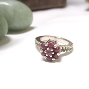 May include: A silver ring featuring a cluster of seven round, deep pink cabochon stones arranged in a floral pattern at the center, with smaller clear stones set along the sides of the polished silver band.