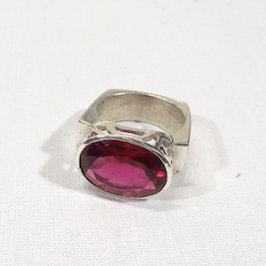 May include: A silver ring featuring a large, oval-cut, deep pink gemstone. The ring has a square band with decorative cutouts near the gemstone setting. The gemstone is the focal point, catching the light and adding a pop of color.