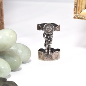 May include: A silver ring featuring a central figure with a sunburst-style headdress and etched details on the band. The figure stands atop a curved, scalloped base, with additional engraved patterns visible on the sides of the metal ring band.
