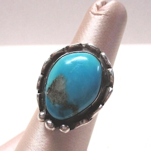May include: A silver ring featuring a large, oval-shaped turquoise stone with a prominent dark matrix inclusion. The metal setting has a textured, notched edge and small spherical accents at the base, framing the vibrant blue-green gemstone.