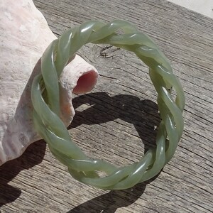May include: A green jade bangle bracelet with a braided design. The bracelet is made of a smooth, polished jade and has a natural, earthy look.